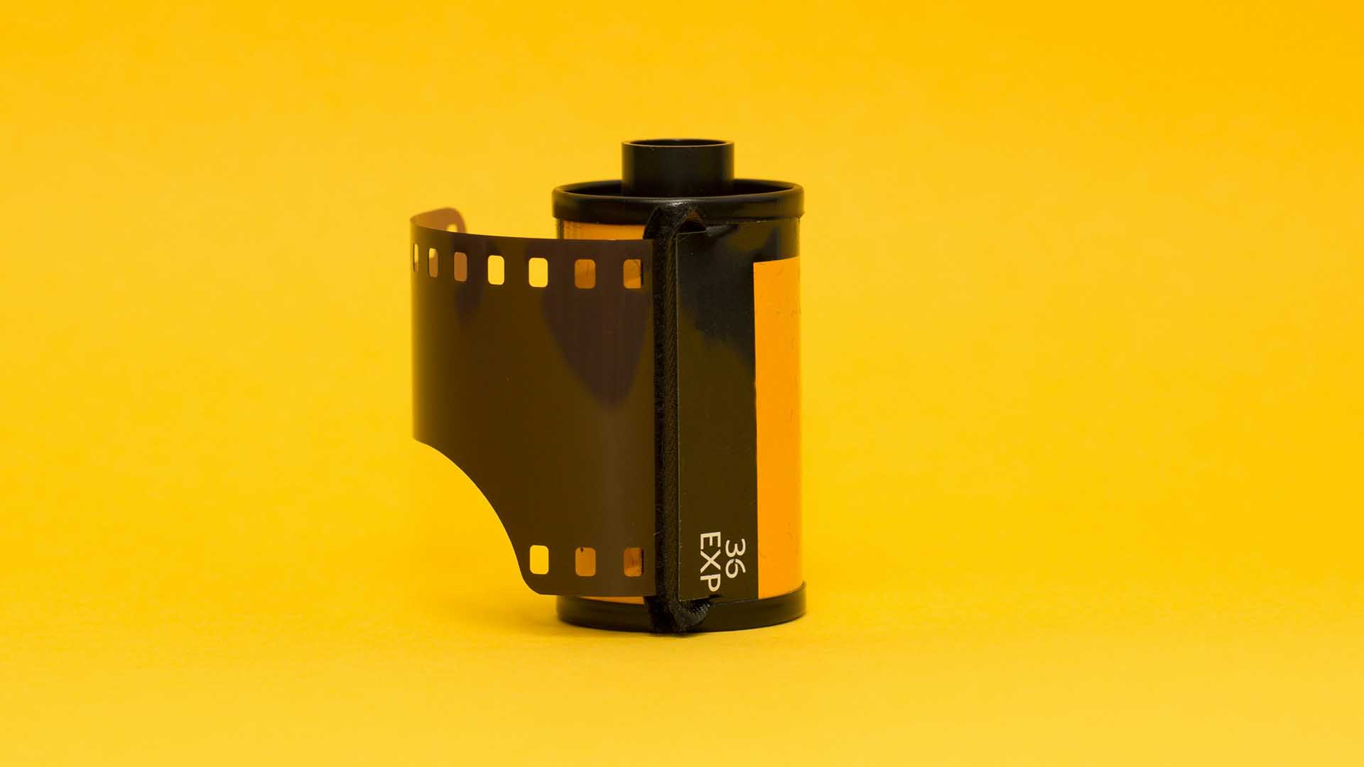 35mm film roll with exposed film leader extending outward, standing upright against a vibrant yellow background.