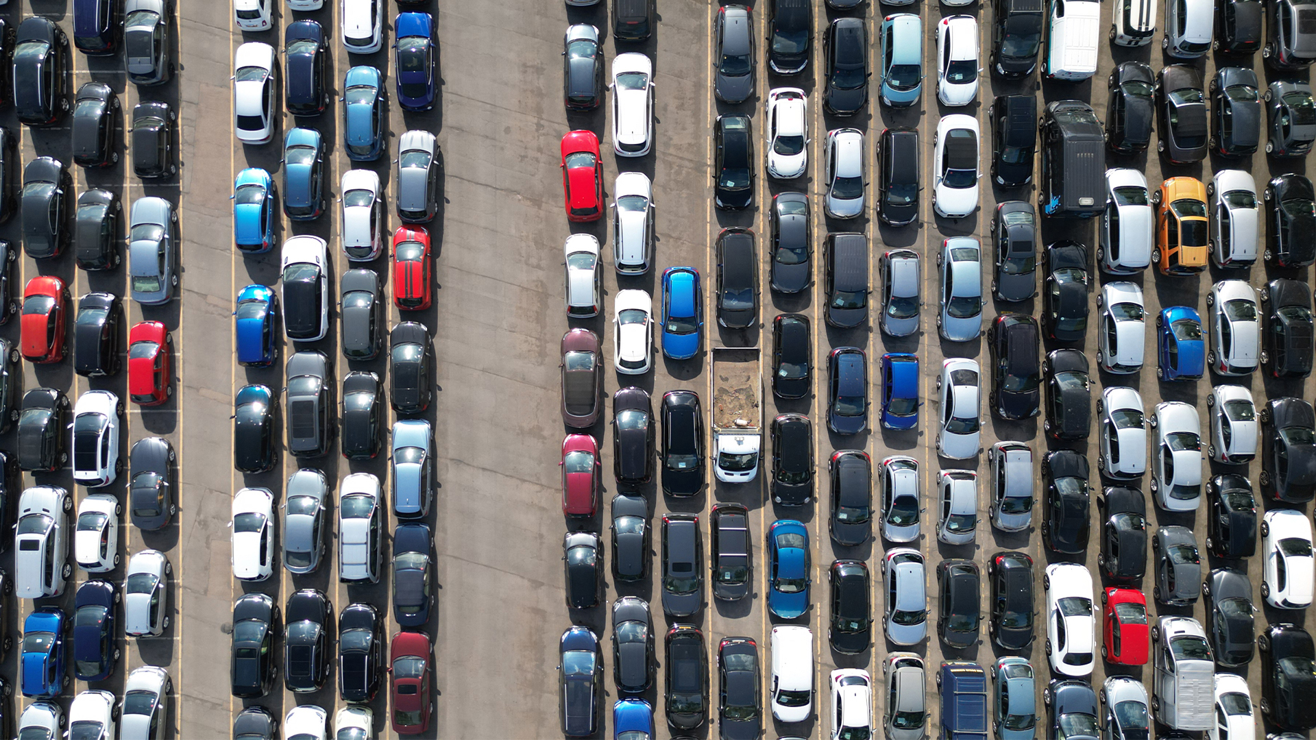 Aerial view of a densely packed parking lot filled with rows of parked cars in various colors and sizes, arranged in parallel lines on a concrete surface.