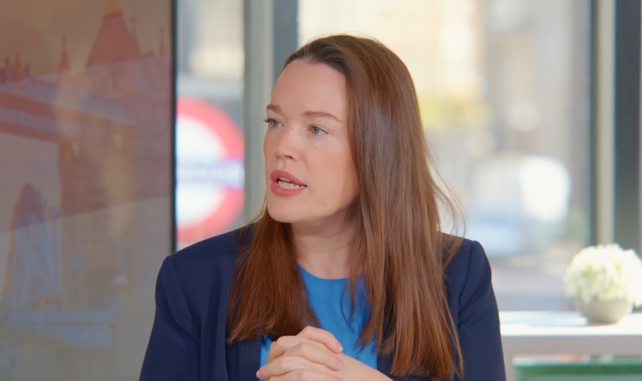 Woman with long brown hair wearing a blue top and dark blazer, speaking with hands clasped, seated indoors with a blurred London Underground sign and cityscape in the background.