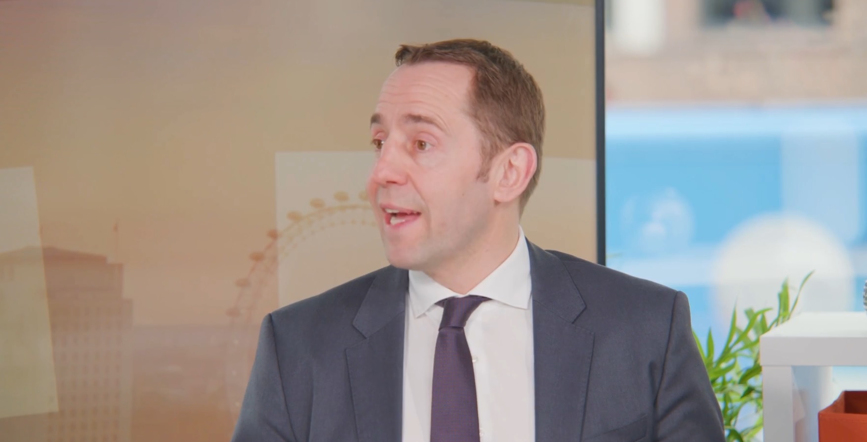 Man in a dark suit, white shirt, and dark tie speaking in a studio setting, with the London Eye faintly visible in the background and a potted plant on a shelf to the right.