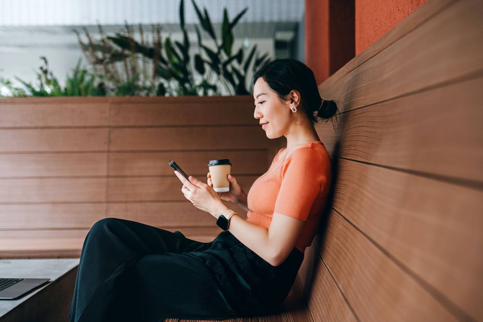 A lady sits on a bench with mobile phone and coffee in hand