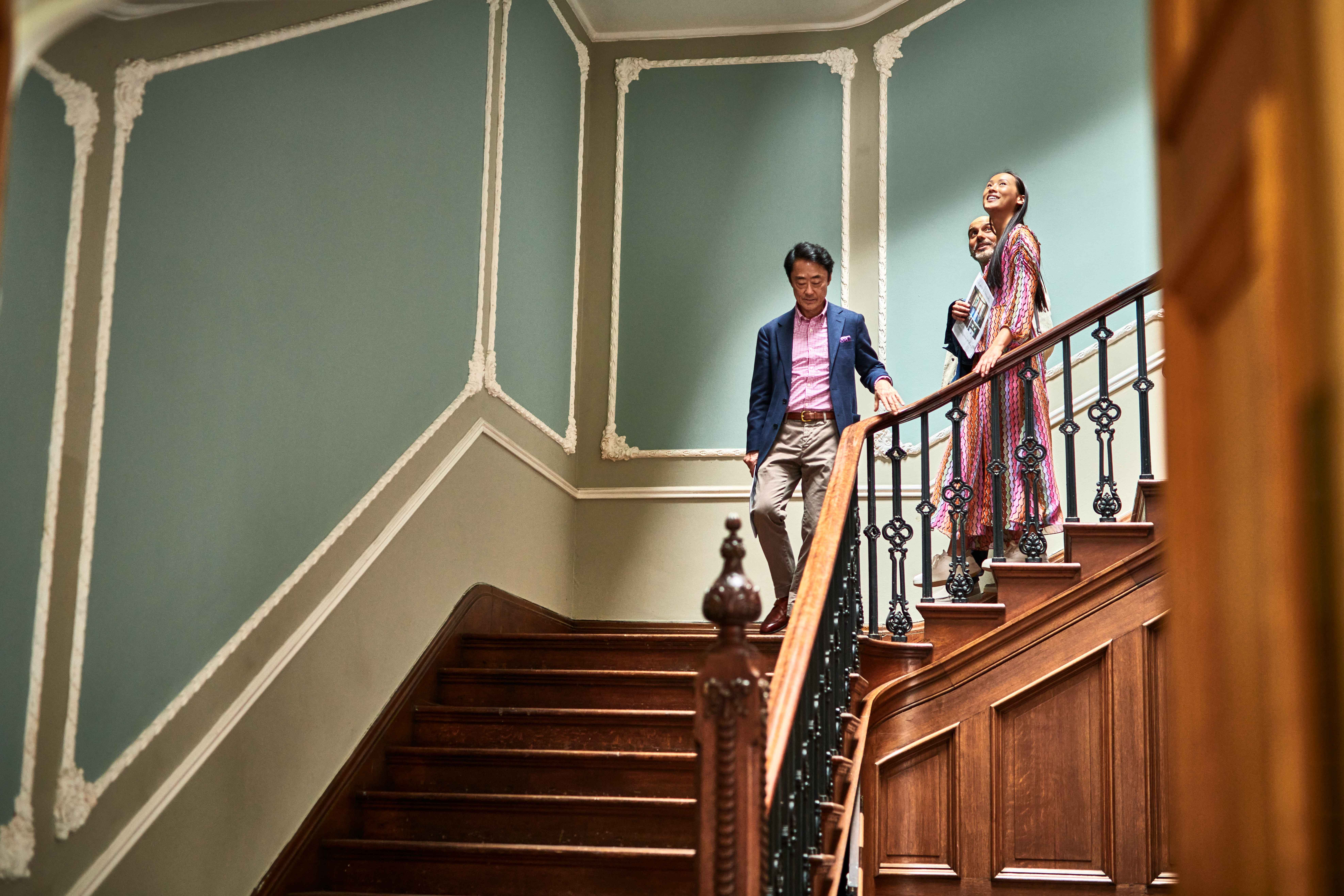A young couple walk down the stairs of a stately home, behind an estate agent