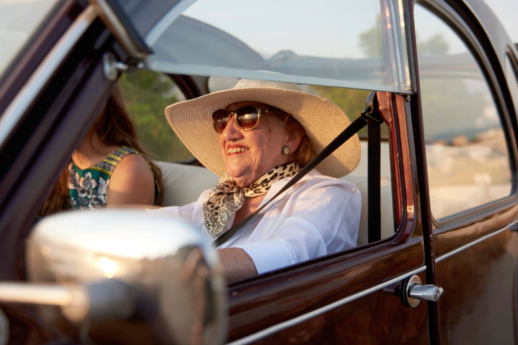 A senior lady wearing sunglasses and a sun hat smiles while driving in a vintage car