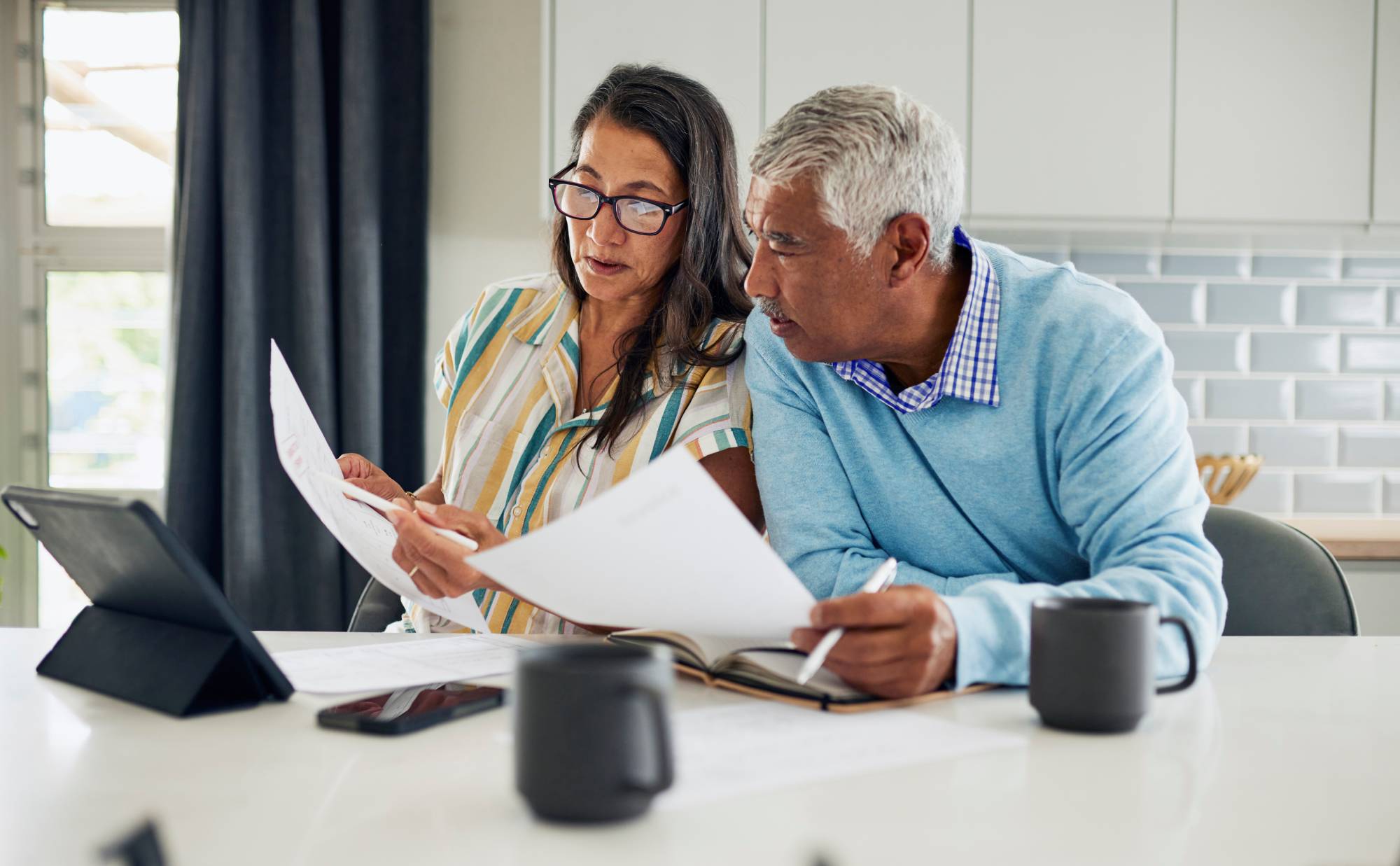 A couple review paperwork at their kitchen bench with coffee and a tablet