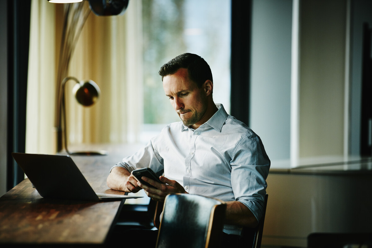 A man with a laptop on a table looks at his mobile phone