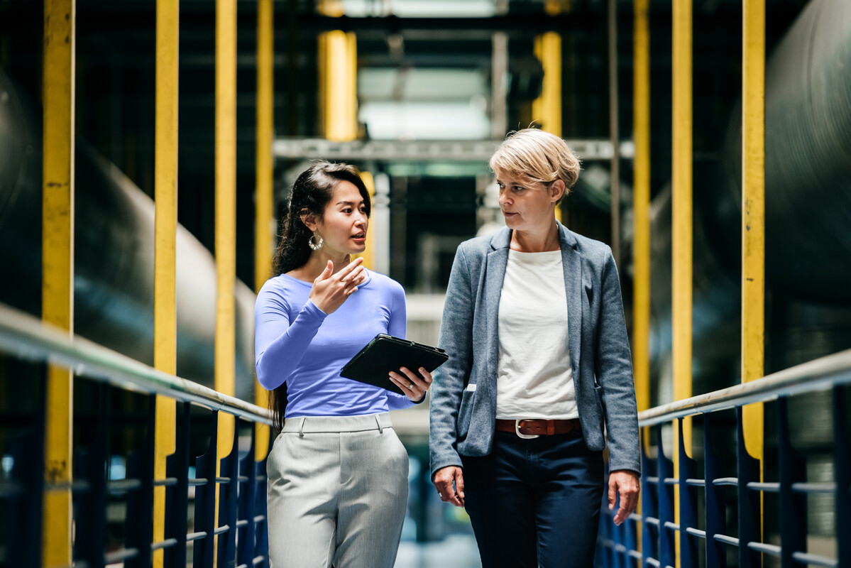 Two businesswomen walk along a footbridge through a factory