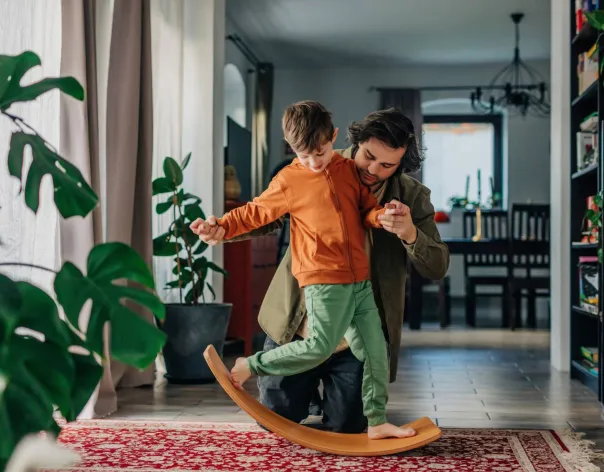 A father helps his son play on a rocking board at home