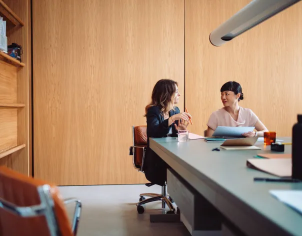 Two women discuss a document together at a large desk in an office