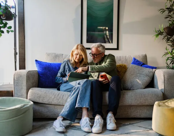 A couple sit together on a grey sofa while looking at a tablet