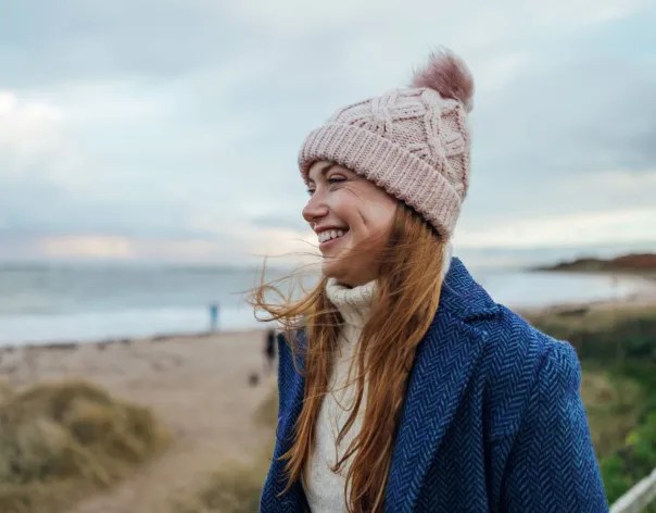 A woman smiles while wearing a beanie at a beach in the winter