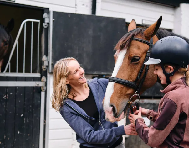 An instructor and young horse rider interact with one another outside some stables
