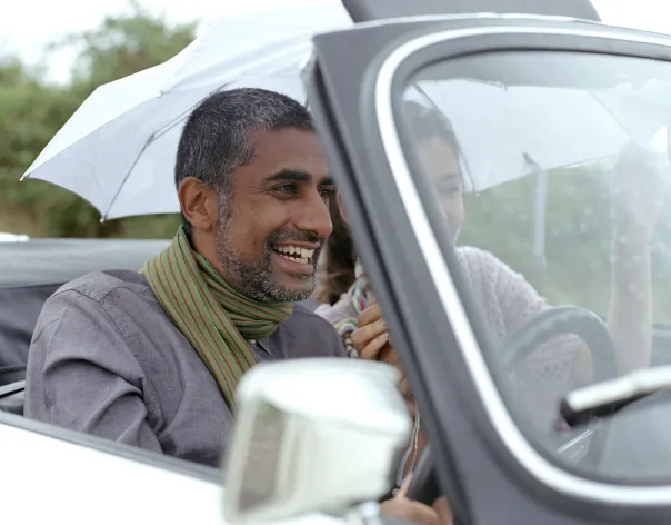 A man and woman laugh together as they drive an old open-topped car while it's spitting rain
