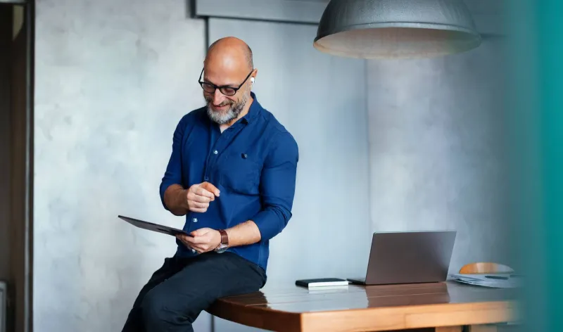 A man sits on the edge of a table while looking at a tablet
