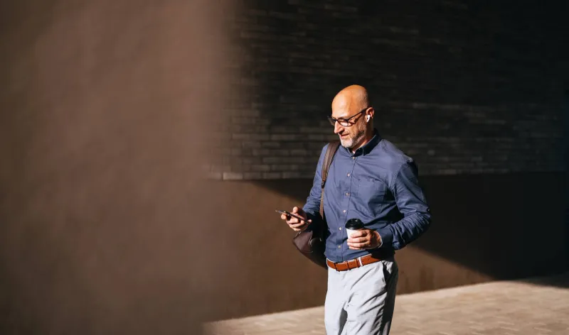 A man walks alongside a building into the sun while using his mobile phone and holding a coffee