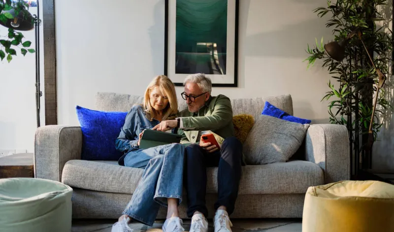 A couple sit together on a grey sofa while looking at a tablet