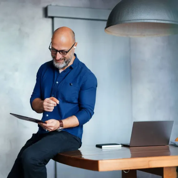 A man sits on the edge of a desk reading a tablet
