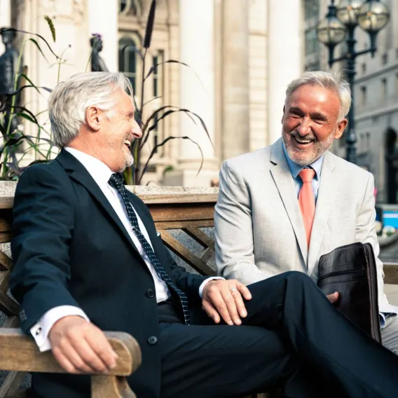 Two men in suits sit on a bench in the city having a conversation