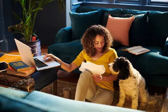 A lady sits on the floor with her dog while working on her laptop