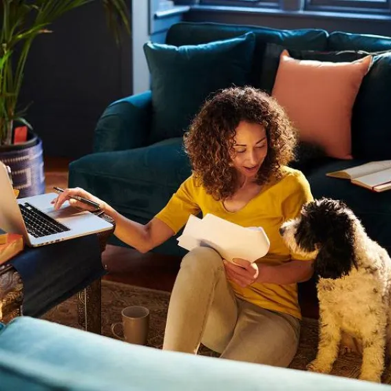A lady sits on the floor of her house with her dog and a laptop