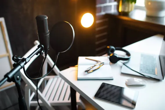 A microphone for recording audio stands in a room that includes a desk with a laptop, headphones and a tablet on it