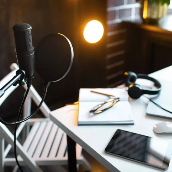 A microphone for recording audio stands in a room that includes a desk with a laptop, headphones and a tablet on it