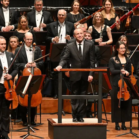The Bournemouth Symphony Orchestra stands while the audience applauds, with conductor Mark Wigglesworth in front