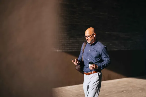 A man walks along a building into the sunlight while holding a phone and a coffee cup