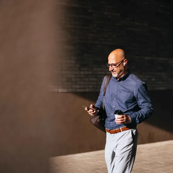 A man walks along a building into the sunlight while holding a phone and a coffee cup