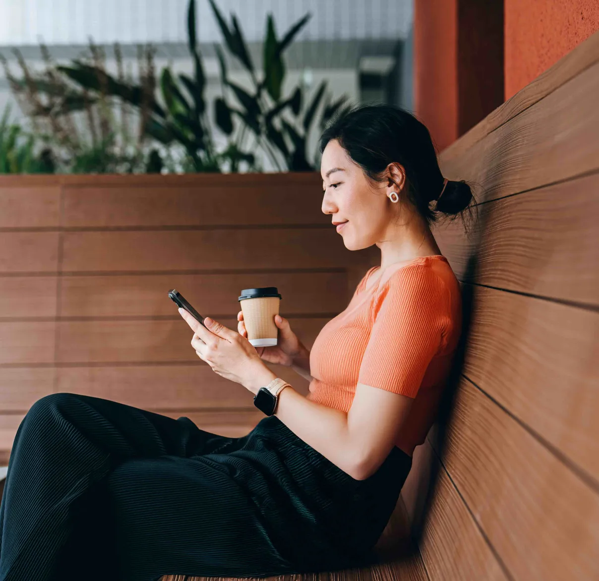 A lady looks at her phone while having a coffee on a bench outside
