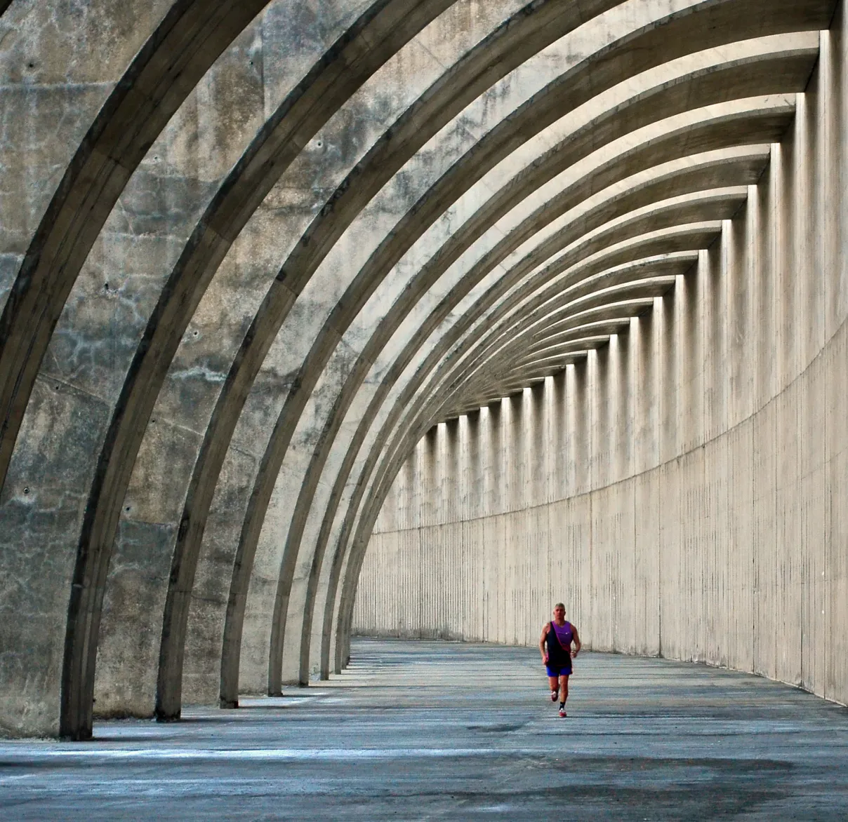 Person jogging through a long, curved concrete corridor with repeating arch-like supports creating a rhythmic pattern of shadows and light.