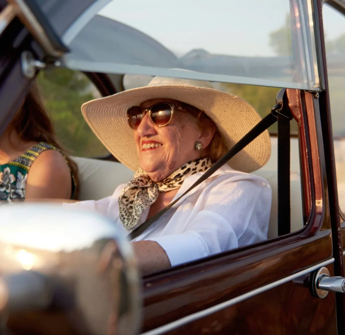 A senior lady wearing sunglasses and a sun hat smiles while driving in a vintage car