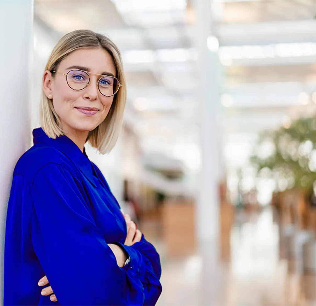 A woman in a blue shirt with crossed arms looks at the camera while standing in an auditorium
