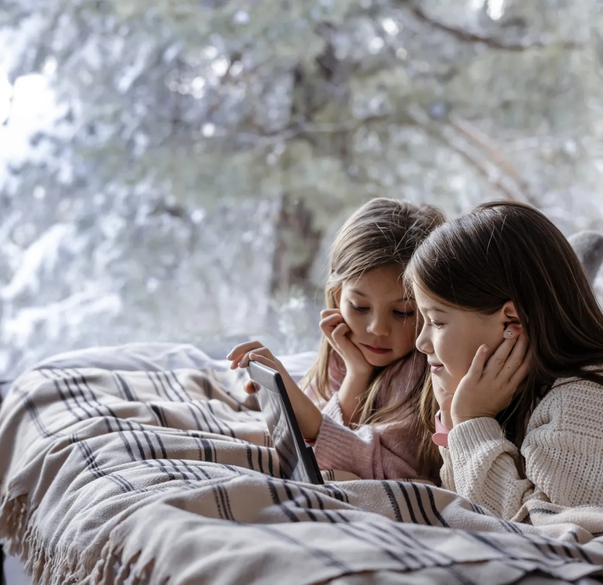 Two children lying under a plaid blanket indoors, watching a tablet near a large window with snowy trees outside