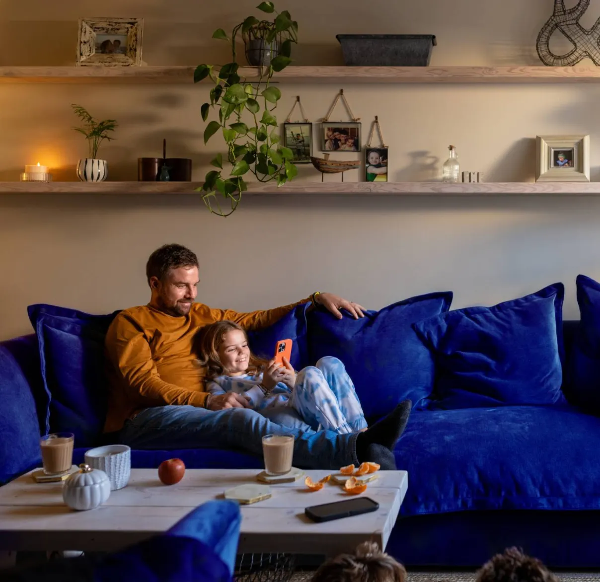 A father and daughter sit together on their sofa at home, while she uses a smartphone