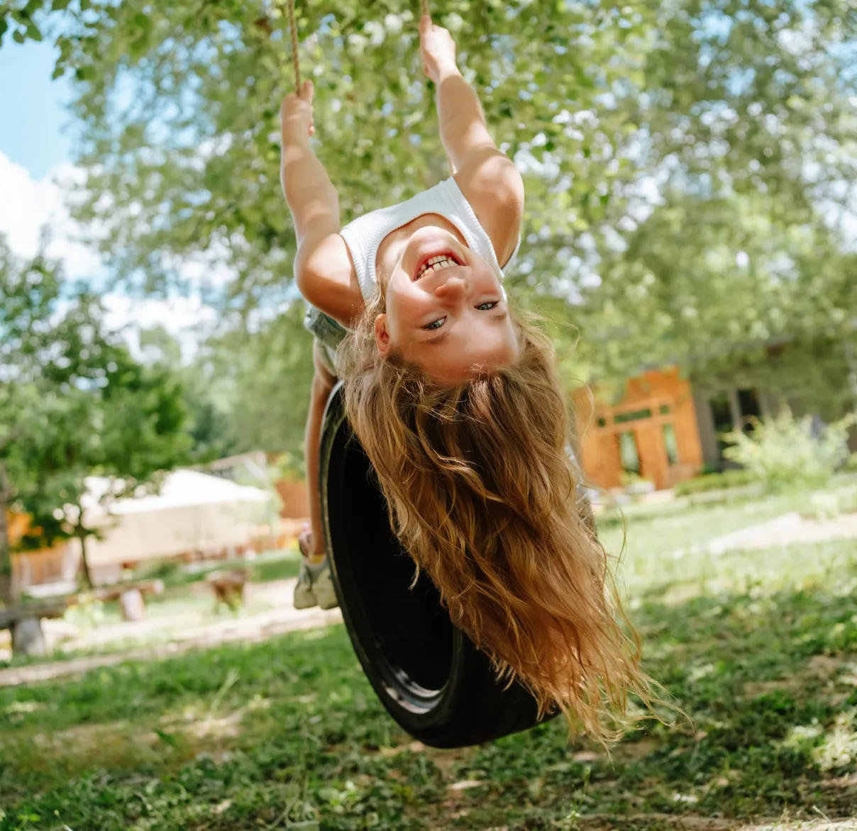 A young girl leans back on a tree swing and smiles while playing in a sunny playground