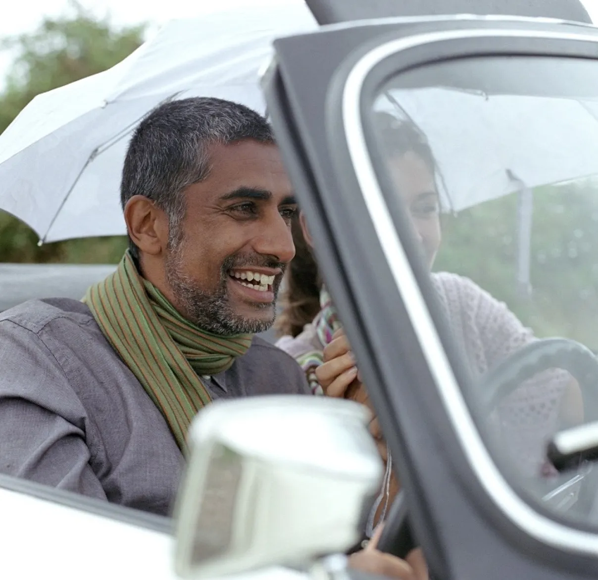 A man and women laugh as they sit in their convertible car under an umbrella as it rains