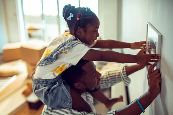 A daughter sits on her father's shoulders and helps to put a photo frame on a wall