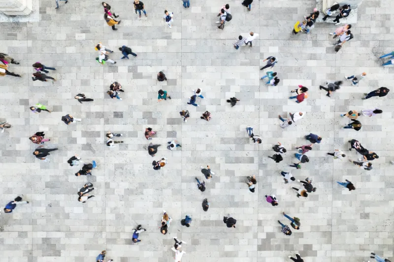 Overhead view of people walking across a large grey stone plaza in different directions, forming a dispersed crowd pattern.