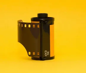 35mm film roll with exposed film leader extending outward, standing upright against a vibrant yellow background.