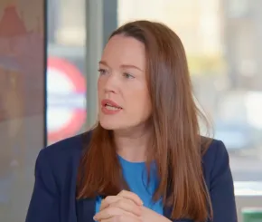 Woman with long brown hair wearing a blue top and dark blazer, speaking with hands clasped, seated indoors with a blurred London Underground sign and cityscape in the background.
