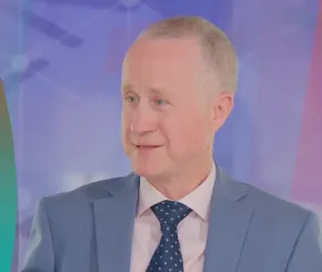 Middle-aged man with short gray hair wearing a light pink shirt, navy polka dot tie, and light blue suit jacket, speaking in front of a colorful abstract background with geometric patterns.