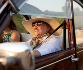 A senior lady wearing sunglasses and a sun hat smiles while driving in a vintage car