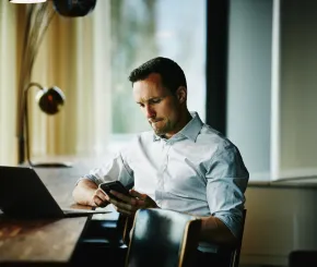 A man with a laptop on a table looks at his mobile phone