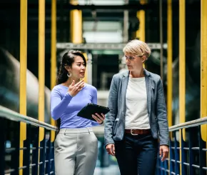 Two businesswomen walk along a footbridge through a factory
