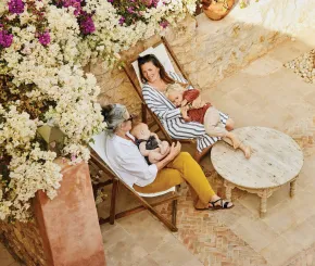 A mother reclines on a deck chair while on holiday, with a child in her arms, while her mother holds another child
