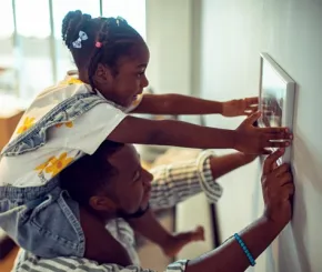 A daughter sits on her father's shoulders and helps to put a photo frame on a wall