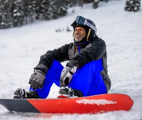 A man takes a break from snowboarding in the mountains to sit on the snow and admire the view