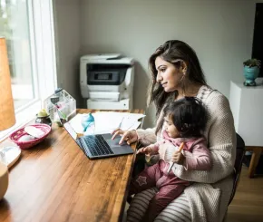 A mother holds her daughter on her lap while she works on a laptop at home