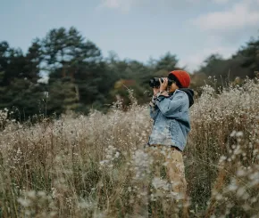 A lady stands in the long grass of a field while looking through binoculars