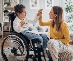 A mother holds the hand of her daughter as she reads a book while sitting in a wheelchair at home